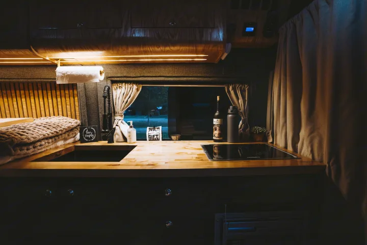 Warmly lit RV rental camper kitchen featuring a wood counter, sink, cooktop, and a window view to the outside.. Dodge 1500 2020