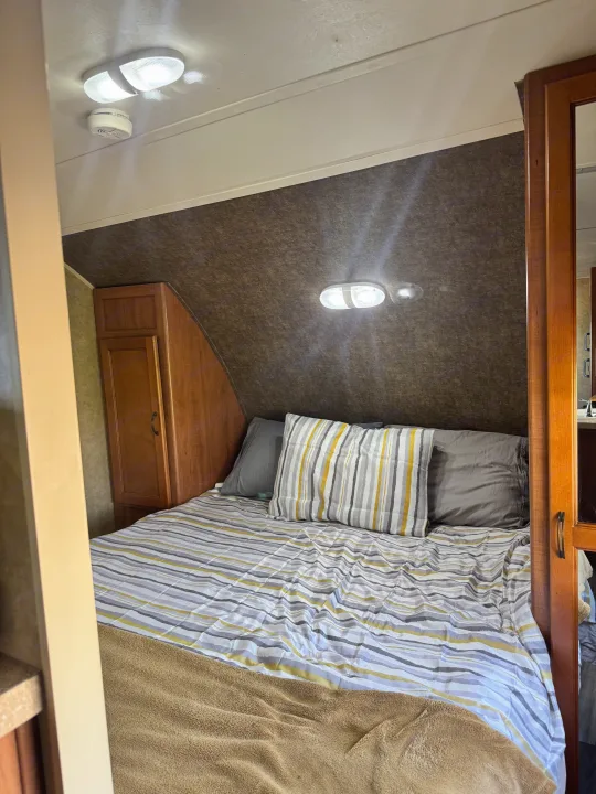 Inside an RV rental: a compact bedroom with striped bedding, a wooden cabinet, and ceiling lights. Ideal for cozy travels.. Forest River Cherokee Grey Wolf 2013