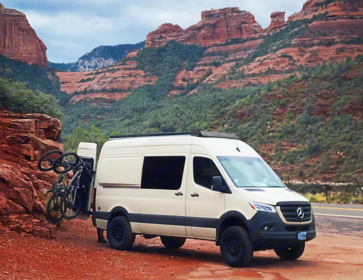 White RV rental camper van with bikes on a red dirt road, set against magnificent red rock mountains.. Mercedes-Benz Sprinter 2020