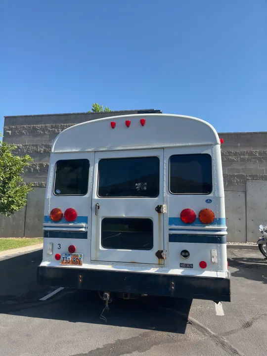 Rear of a white short bus, a potential rv rental, parked in a sunny lot. Features Utah plate, red lights, & rust on hinges.. Chevrolet G3500 Express 2006