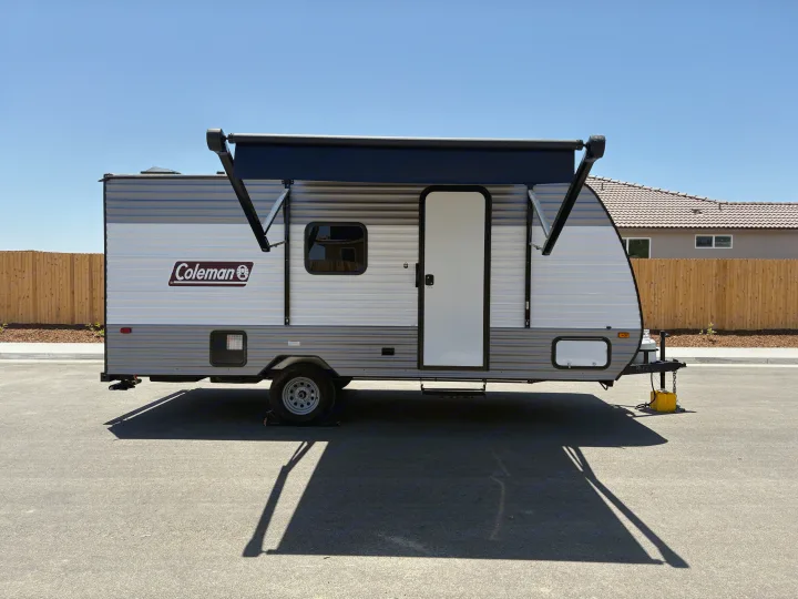 A striped white and grey travel trailer with a black extended awning, parked on asphalt, ready for rv rental.. Dutchmen Coleman 2024