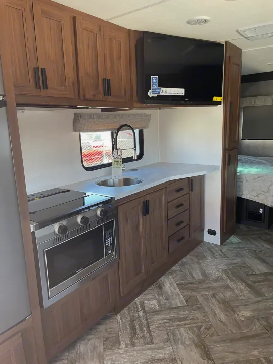 Modern RV rental interior with a compact kitchen: sink, microwave, stove, TV, and wood-look flooring. Bedroom visible.. Forest River Sunseeker 2022
