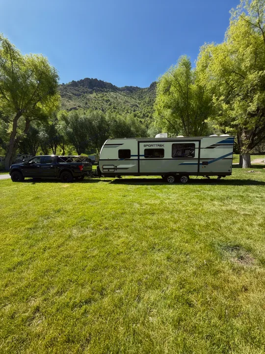 RV rental setup: A black truck tows a travel trailer on lush green grass, with scenic trees and mountains.. Sport Trek 270 VBH 2021