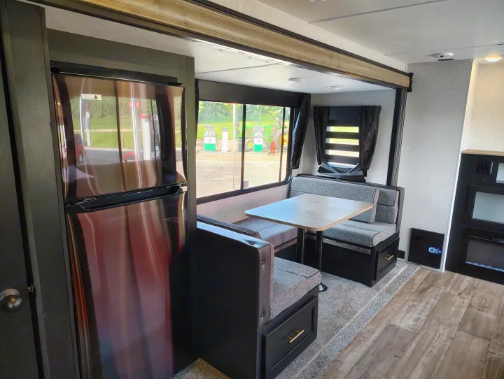Inside an RV rental: a modern dinette, refrigerator, and window view of a gas station and tractor outside.. Forest River Patriot 274BRB 2022