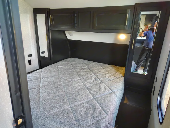 Inside an RV rental: a compact bedroom with dark cabinets, gray quilted bed, built-in light, and mirror.. Forest River Patriot 274BRB 2022