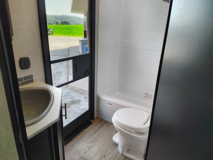 Tour an RV rental's efficient bathroom: sink, toilet, and shower/tub unit. A screen door reveals scenic fields.. Forest River Patriot 274BRB 2022