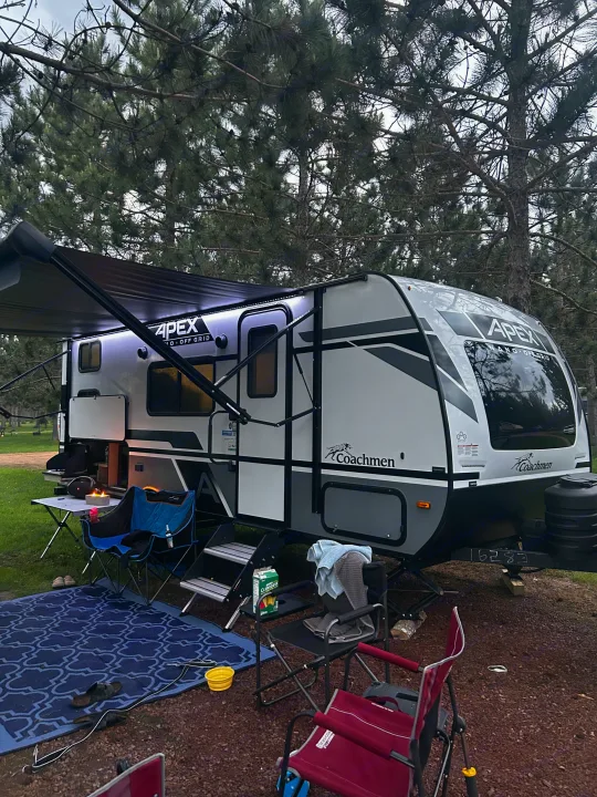 RV rental set up for camping. An Apex travel trailer with an illuminated awning, outdoor area, and pine trees.. Coachmen Apex Nano 2023