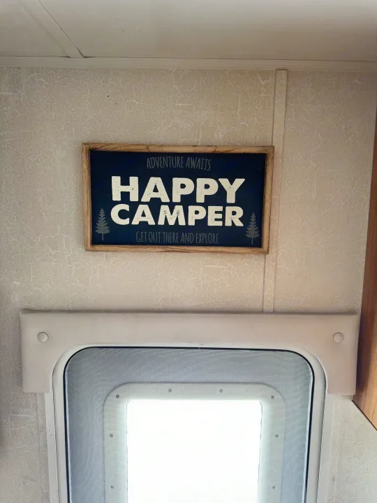 RV rental interior view: A 'Happy Camper' sign on a textured wall, positioned above a bright screen door.. Coachmen Leprechaun 2001