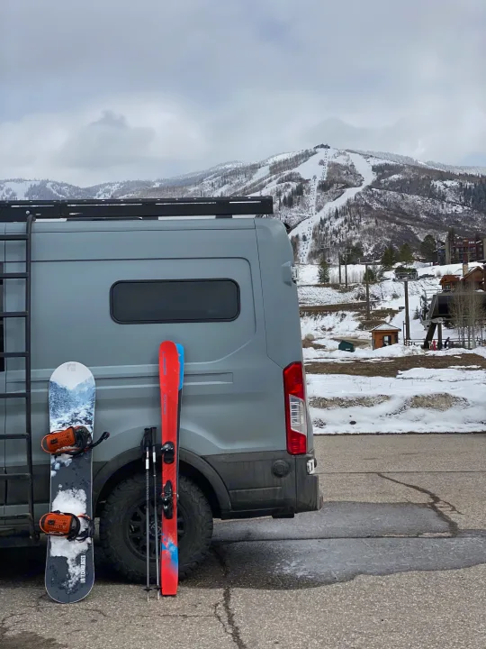 RV rental ready for ski day. Grey van with snowboard & skis, snow-covered mountains, & ski lifts in background.. Ford Transit 2019