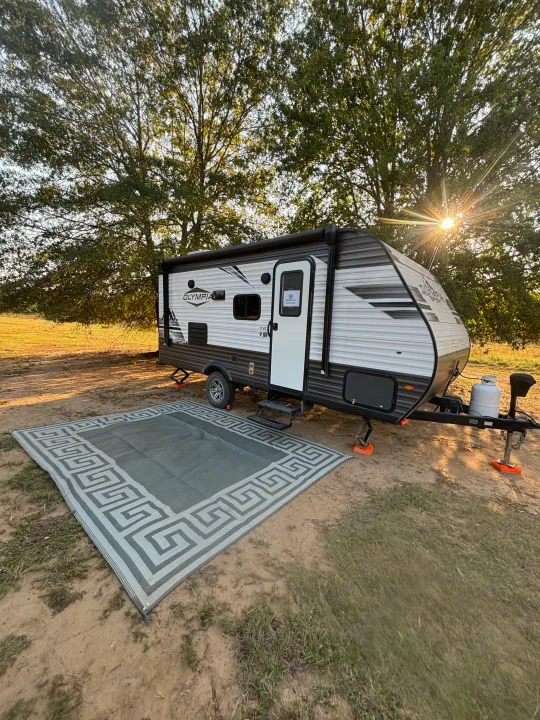 Sunny RV rental setup: a travel trailer with an outdoor rug on a spacious campsite surrounded by trees.. Olympia Sport 19BH 2022