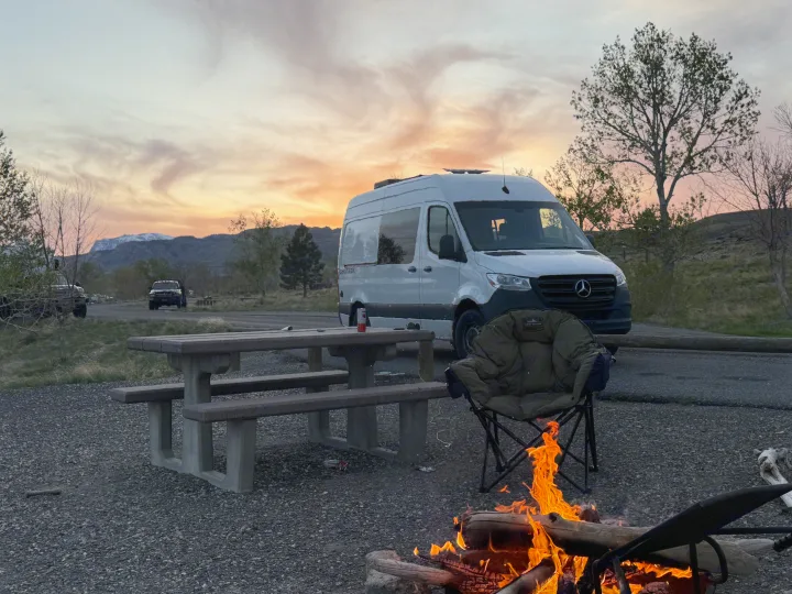 Enjoying the golden hour by a warm campfire at a scenic campsite with a white RV rental van, perfect for outdoor adventures.. Mercedes-Benz Sprinter 2024