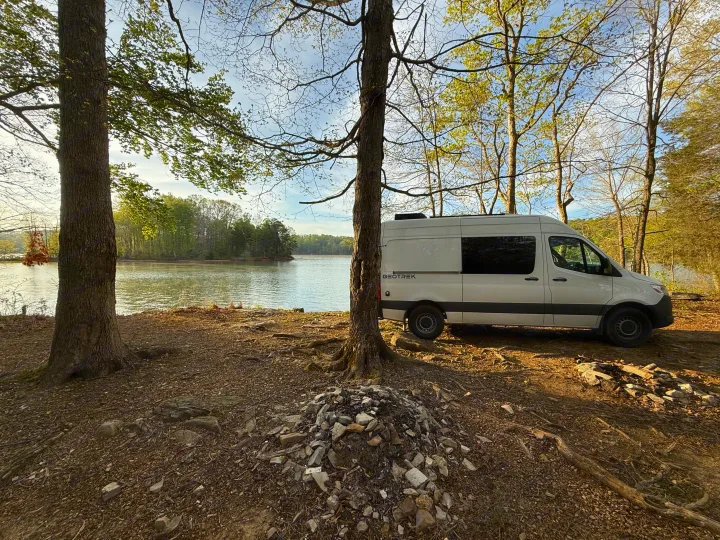 Lakeside RV rental: A white camper van parked on a dirt campsite surrounded by trees, offering peaceful nature views.. Mercedes-Benz Sprinter 2024