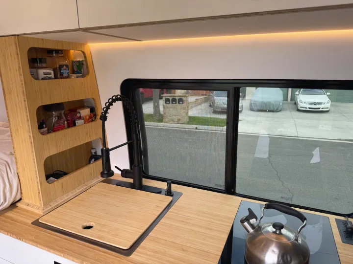 This RV rental's modern bamboo kitchen offers a sink, stovetop, and organized storage, with a view outside.. Mercedes-Benz Sprinter 2024