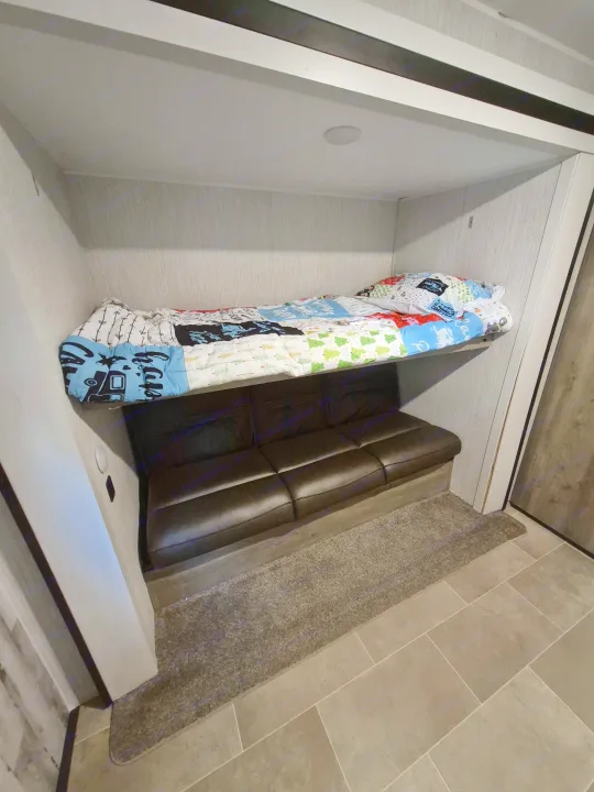 Compact RV rental space with a cozy bunk bed above a sofa. Features patterned quilt, built-in light, and mixed flooring.. Forest River Palomino Puma XLE Lite 2021