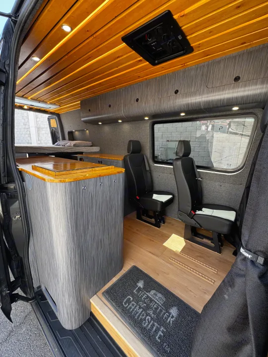 Modern RV rental interior: wood slat ceiling, gray cabinets, bed, kitchen counter, & passenger seats. Ready for adventure.. Mercedes-Benz Sprinter 2023