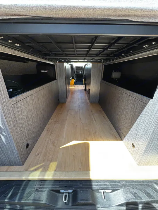 Spacious RV rental interior with wood floor, custom storage, and overhead frame for comfortable mobile living.. Mercedes-Benz Sprinter 2023
