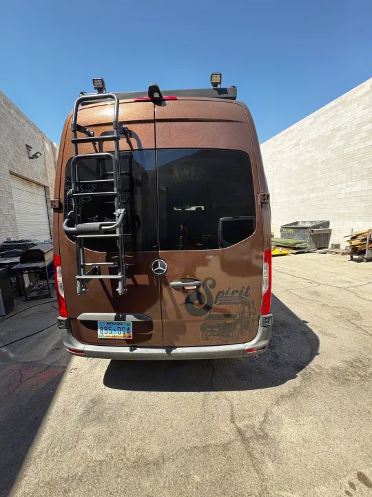 Rear of a brown Mercedes-Benz Sprinter RV rental van, customized with a ladder, roof lights, & 'Spirit' graphic. Parked outdoors.. Mercedes-Benz Sprinter 2023