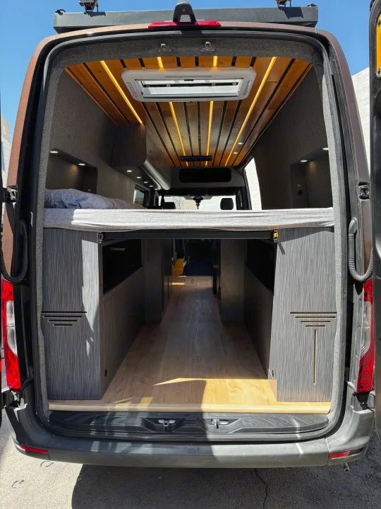 Explore this modern RV rental's custom camper van interior, showcasing a raised bed, wood ceiling, ambient lighting, and storage.. Mercedes-Benz Sprinter 2023