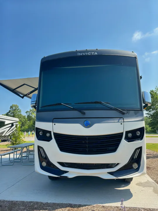Front view of a white and grey Invicta RV rental, parked with its awning out at a campsite with a picnic table.. Holiday Rambler Invicta 2021