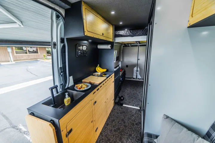 Inside an RV rental: a modern camper van kitchen with sink, food prep area, and overhead bed. Ready for road trips!. Mercedes Sprinter 2023