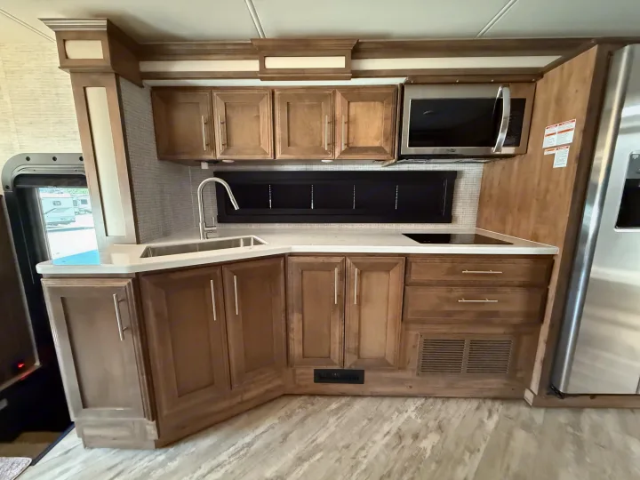 Modern **RV rental** kitchen with light wood cabinets, white countertops, stainless steel sink, microwave, and electric cooktop.. Jayco Seneca 2025