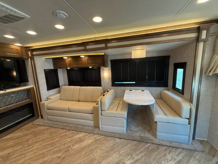 Spacious RV rental interior featuring cream sofas, a dining booth, TV, fireplace, and bright overhead lighting for comfortable travel.. Jayco Seneca 2025