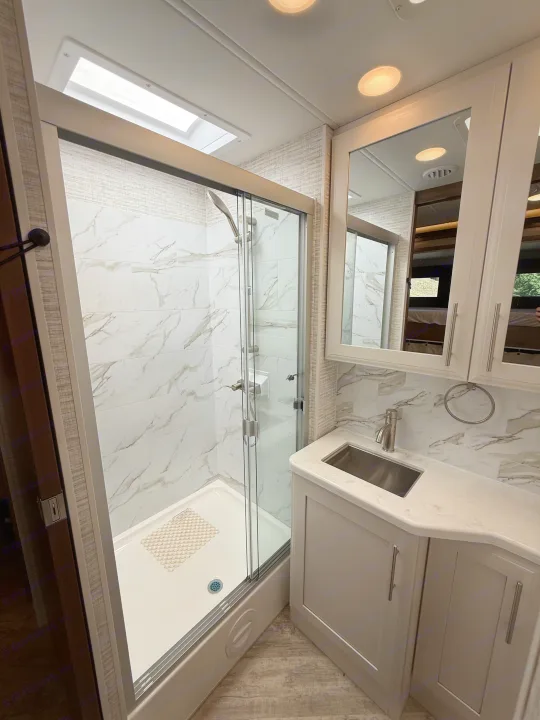 Modern RV rental bathroom features a sleek glass shower with white marble-look tile, skylight, and contemporary vanity with an undermount sink.. Jayco Seneca 2025