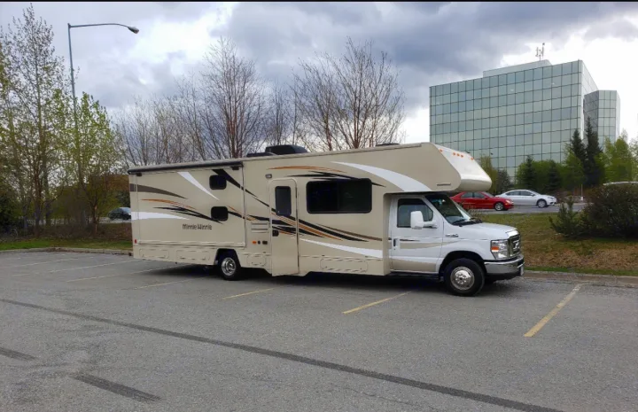 An RV rental, a beige and white Minnie Winnie, is parked in a lot with a glass building and trees in the background.. Winnebago Minnie Winnie 2017