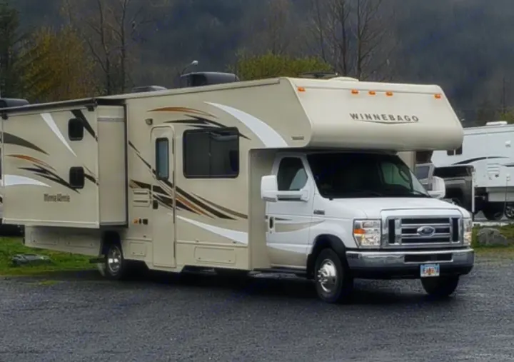 A large, tan Winnebago Minnie Winnie Class C RV rental with an extended slide-out is parked on gravel amidst trees and mountains.. Winnebago Minnie Winnie 2017