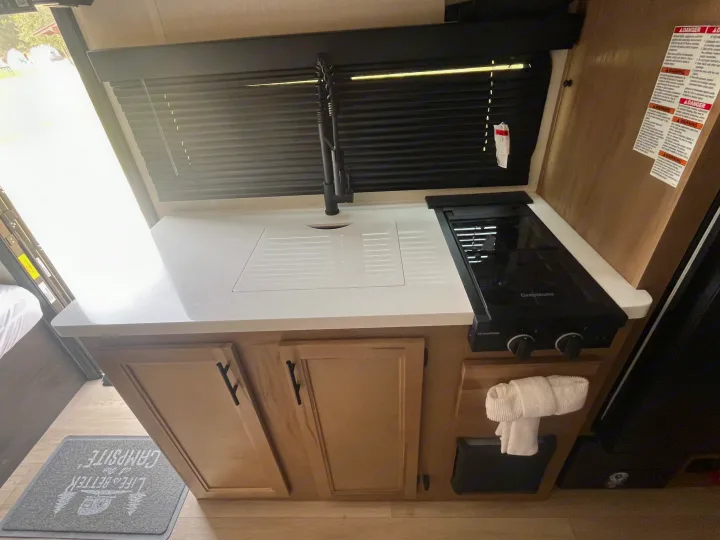 RV rental: Bright, compact kitchen with sink, two-burner stove, light wood cabinets, and window overlooking a campsite.. Forest River Cherokee Cherokee Black Label 2025