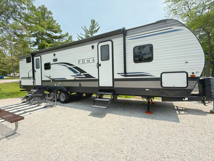 A white Puma RV rental, a travel trailer, set up at a gravel campsite with chairs, mat, and a picnic table.. Forest River Palomino Puma 2022