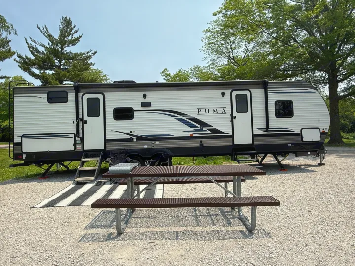 A white Puma travel trailer (rv rental) parked on gravel at a sunny campsite with a picnic table and striped mat.. Forest River Palomino Puma 2022