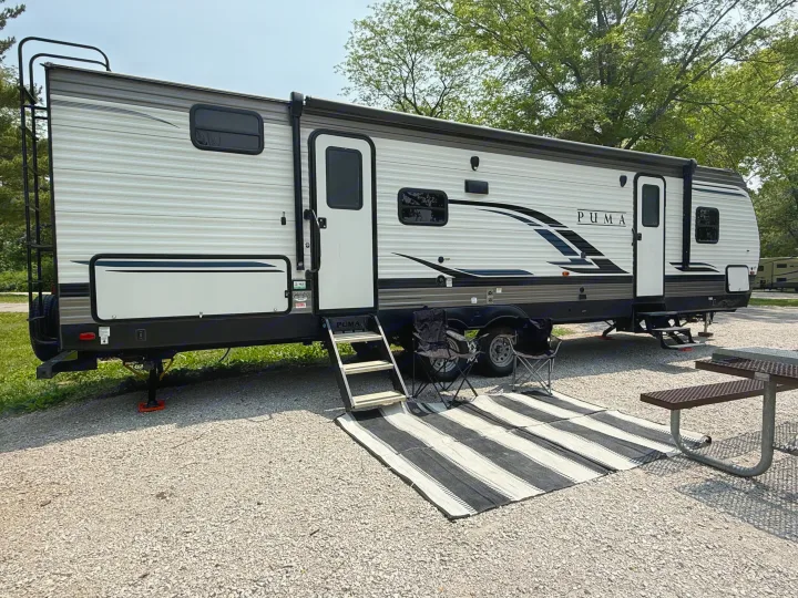 Puma RV rental set up at a gravel campsite with an outdoor rug, chairs, and a picnic table, perfect for camping.. Forest River Palomino Puma 2022