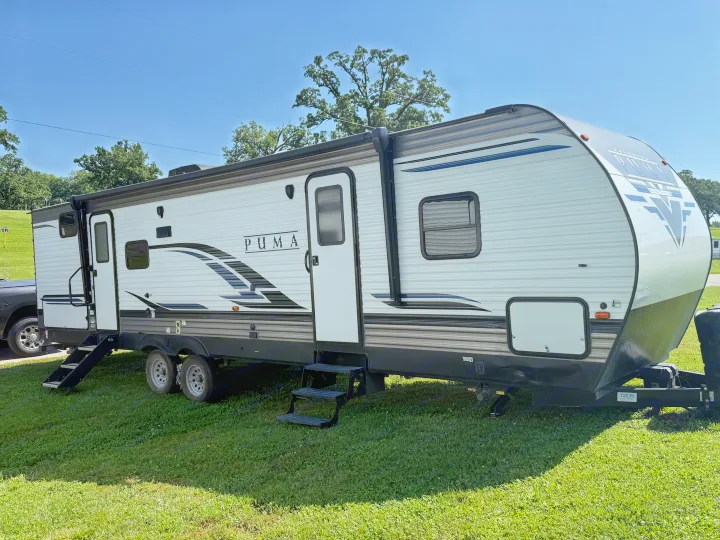 A white and grey PUMA travel trailer RV rental, featuring a large slide-out, parked on green grass under a clear blue sky.. Forest River Palomino Puma 2022