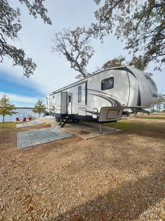 A white and black fifth-wheel RV rental is parked on a concrete pad at a scenic lakeside campsite, with red chairs and gravel.. Forest River Sabre 2022