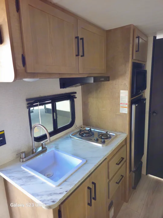 Compact RV rental kitchen with light wood cabinets, sink, two-burner cooktop, microwave, and refrigerator, maximizing space.. Keystone RV Springdale 2025