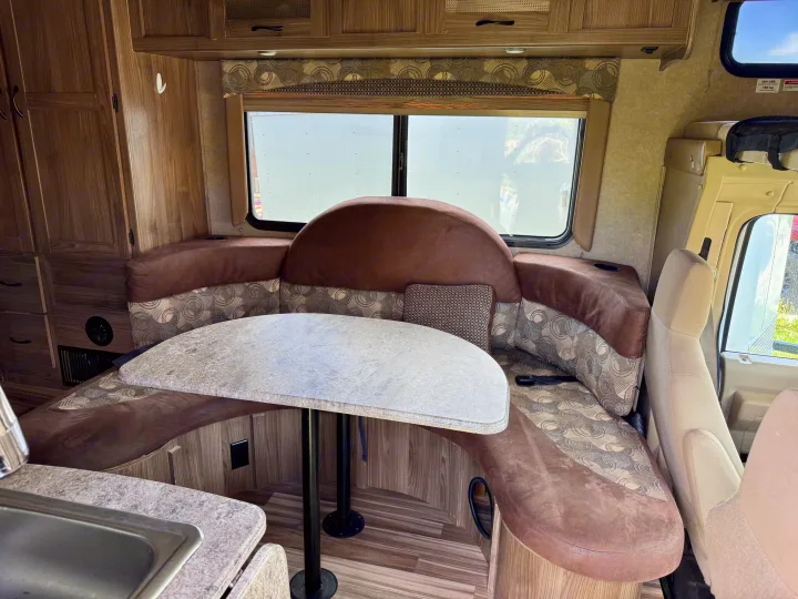 Spacious RV rental dinette with U-shaped booth seating, table, wood-grain cabinetry, and cockpit view.. Coachmen Freelander 21QB 2016