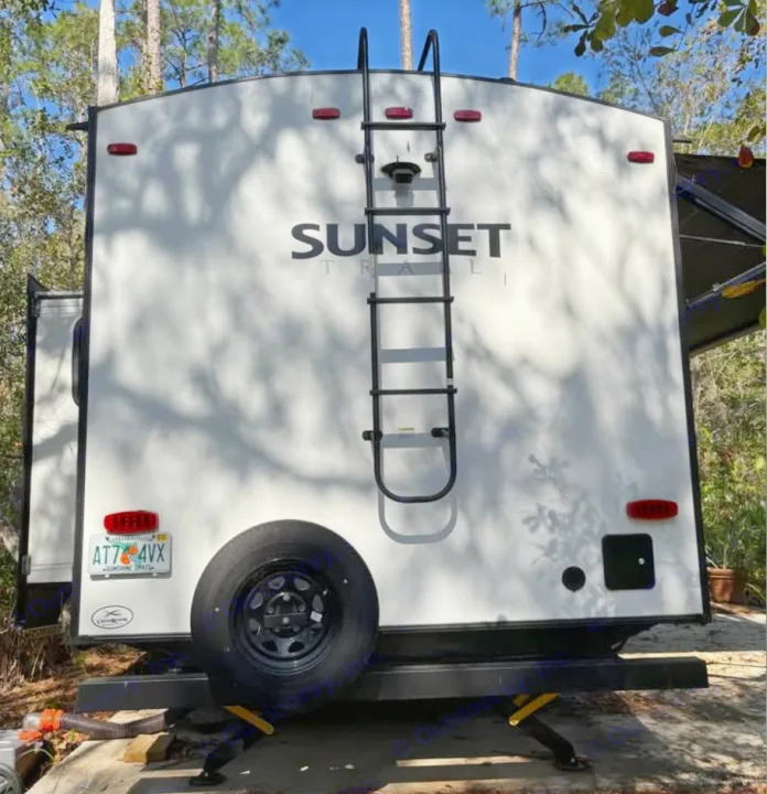Rear view of an RV rental, a Sunset Trail travel trailer, with a roof ladder and spare tire, set up at a sunny campsite.. Crossroads RV Sunset Trail Super Lite 2022
