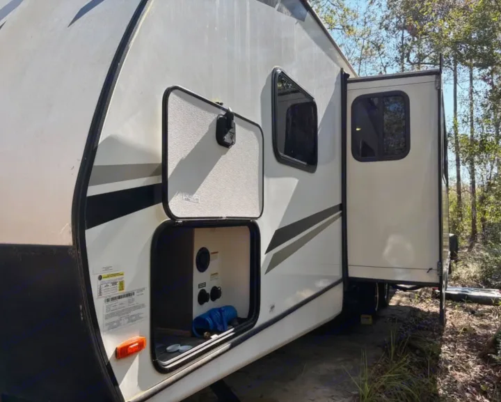This RV rental is parked at a wooded campsite, with its utility storage compartment open and a slide-out extended.. Crossroads RV Sunset Trail Super Lite 2022