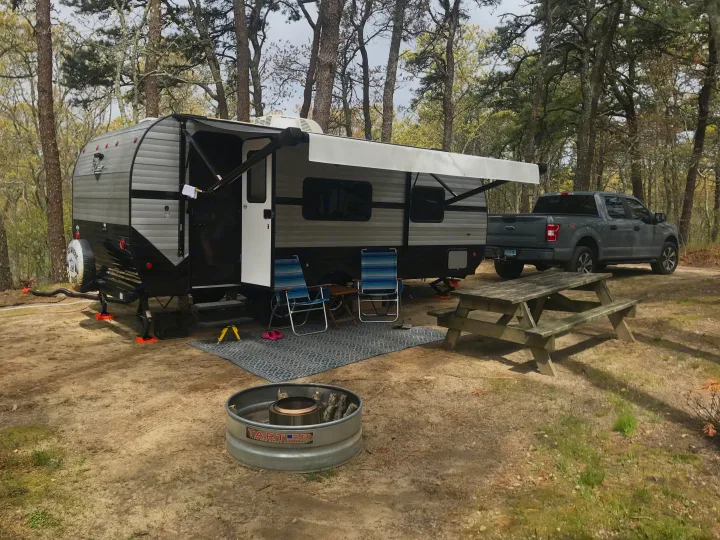 An RV rental, a grey travel trailer, set up at a wooded campsite with an awning, outdoor rug, chairs, and fire pit.. Riverside RV Retro 2021