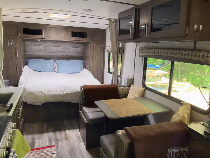 Inside a comfy RV rental: bedroom with a bed, a dining booth, and a kitchen area. Windows offer views of a serene outdoor campsite.. Riverside RV Retro 2021