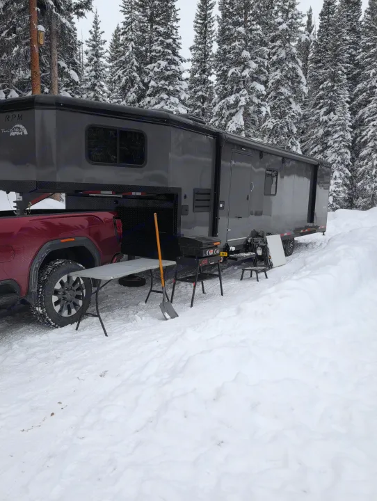 RV rental adventure! A red pickup pulls a dark toy hauler, set for winter camping in a snow-covered forest with outdoor gear.. Trails West RPM Toy Hauler 2023