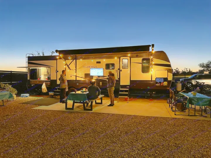 A cozy RV rental evening. People gather outside a travel trailer with string lights, enjoying TV and outdoor camping fun.. Keystone RV Hideout 2021