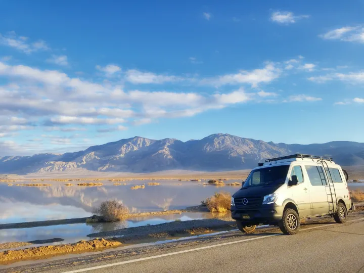 A white adventure rv rental van, likely a Sprinter, is parked by a reflective lake in a mountainous desert. Ideal for road trips!. Sportsmobile Regular Body 4X4 2017