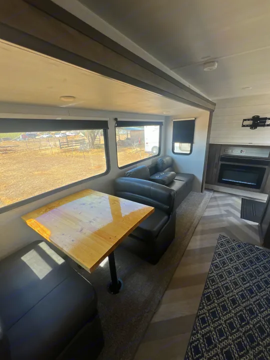 Modern RV rental interior: dining area, black sofa, fireplace, and large windows revealing an outdoor scene.. Forest River Wildwood 2020