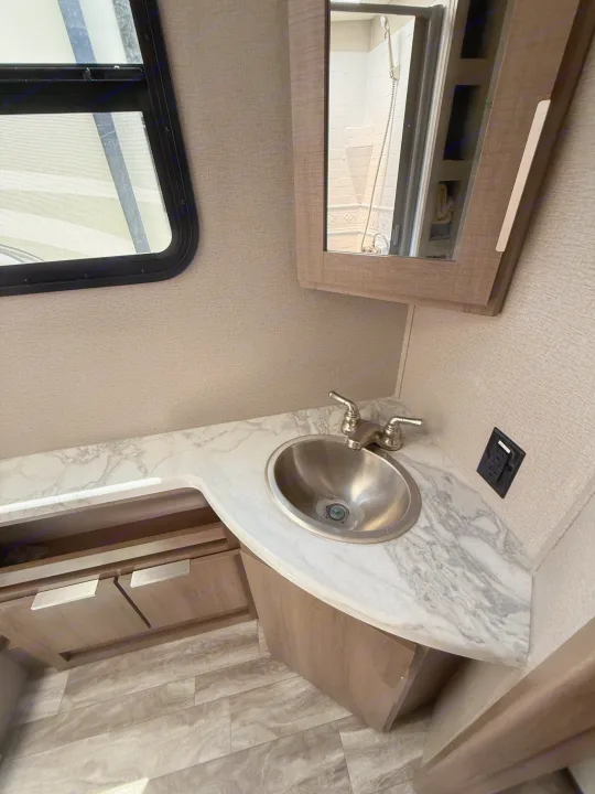 RV rental bathroom with a compact vanity, stainless steel sink, faucet, and mirrored medicine cabinet. A window is on the left.. Grand Design Imagine 2021