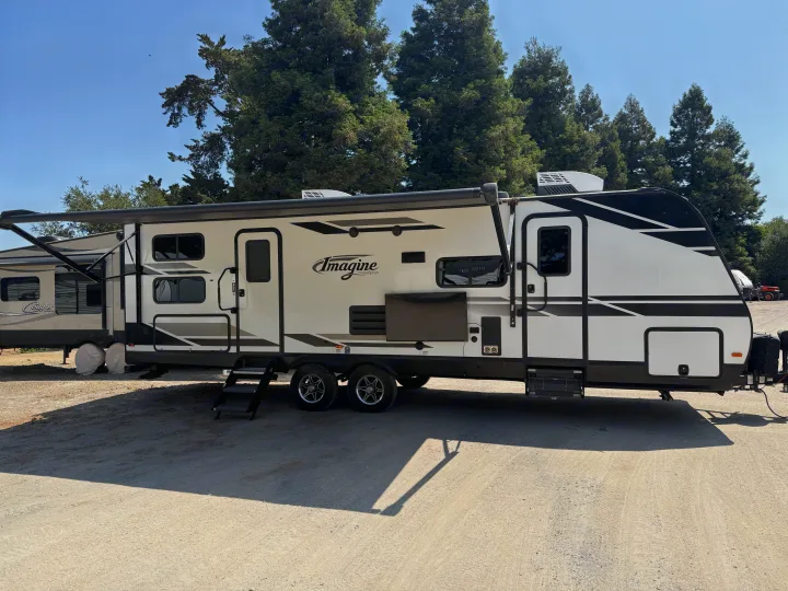 White Imagine travel trailer RV rental, with an extended awning and steps, parked on gravel under a clear sky with trees.. Grand Design Imagine 2021