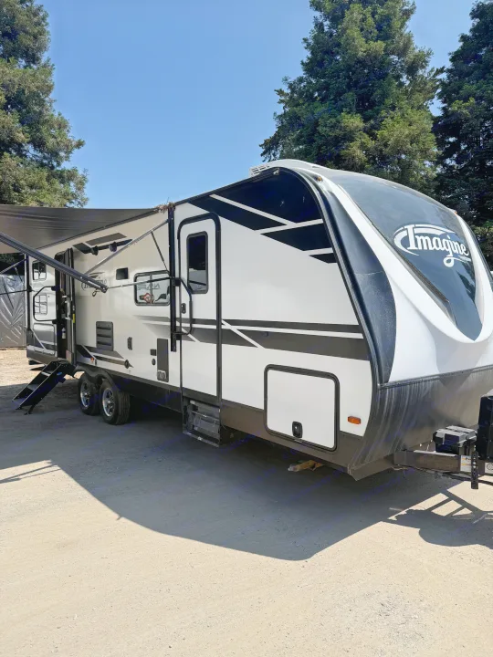 An Imagine travel trailer, white and black, is parked on dirt under a blue sky with trees, ideal for an RV rental.. Grand Design Imagine 2021