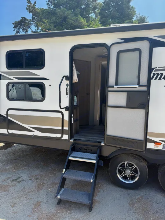 An RV rental, a white travel trailer, with its main door open, revealing steps to the interior. Parked outdoors under trees.. Grand Design Imagine 2021