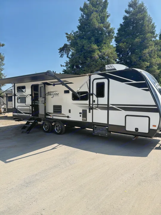 A white and black Imagine travel trailer RV rental, awning extended, steps down, parked on a gravel lot.. Grand Design Imagine 2021